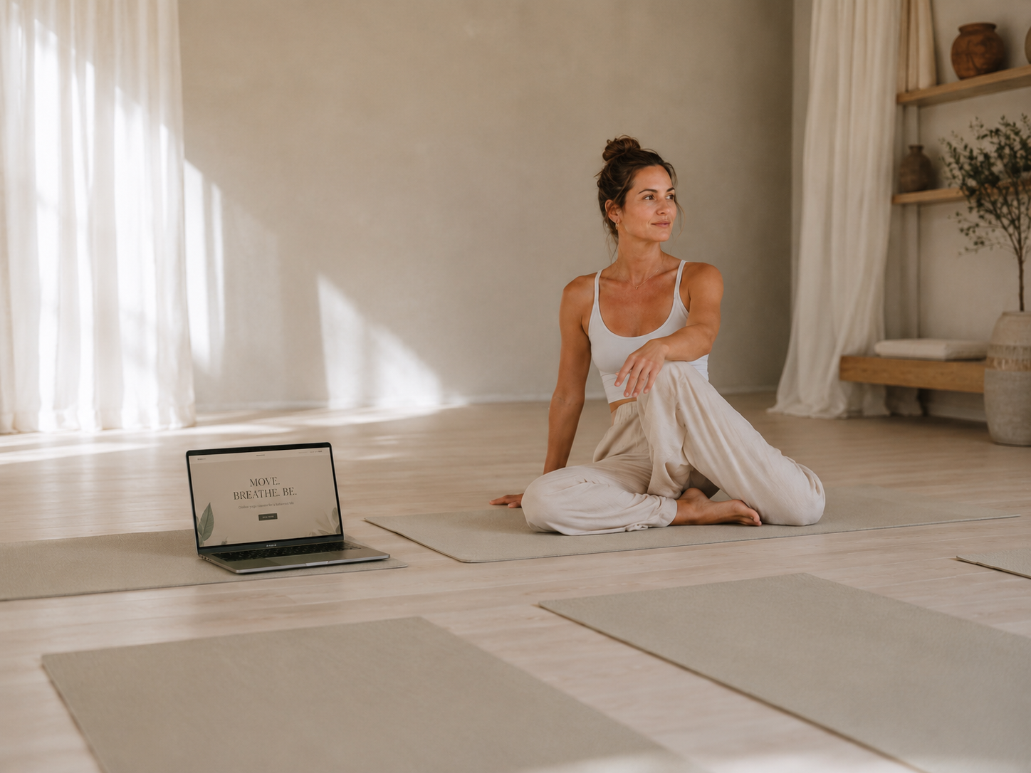 Yoga teacher on her mat in a light-filled studio, with a laptop nearby and empty mats waiting for students